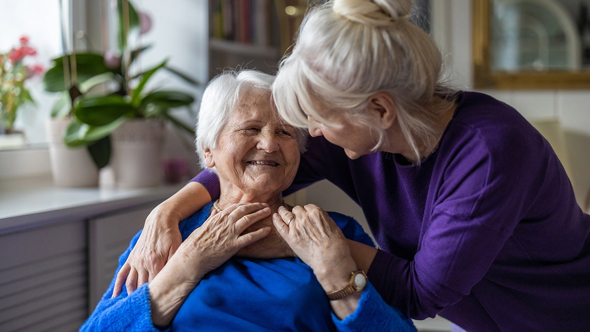 One woman leans down to hug another, older woman from behind the seated woman's shoulder. Both women are older with white hair.