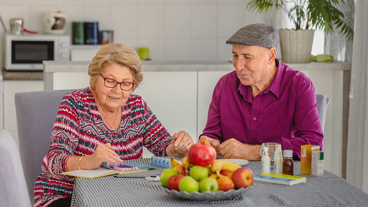 An older woman and older man sit at a kitchen table with a plate of fresh fruit in front of them.