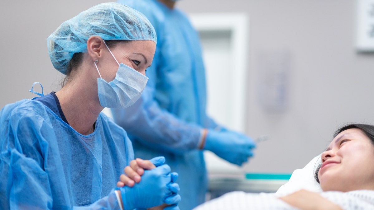 A nurse in blue scrubs holds a pregnant woman's hand in a hospital room.